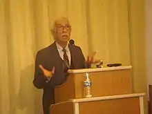 Photographie en couleur d'un homme âgé s'exprimant devant un pupitre et un micro.