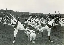 Photographie montrant des gymnastes en exercice, soulevant une barre entre les bras, une jambe relevée.