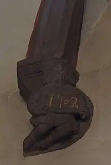 photographie en couleur montrant un culot en grès rose sculpté avec un personnage barbu et vêtu d’habits médiévaux, qui tient une banderole inscrite avec la date 1504