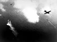 Photographie en noir et blanc prise d'un avion à moyenne altitude. Un autre avion précède celui d'où a été pris la photo et un navire est visible.