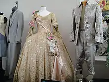 Une robe beige très ornementée, entre autres de grosses fleurs, et un costume gris masculin en satin lui aussi décoré d'un motif floral imprimé. Les deux sont exposés dans une galerie.