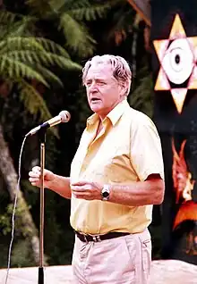 Photographie en couleurs d'un homme portant une chemise jaune parlant debout devant un micro, sur une scène.