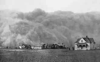 Photographie en noir en blanc montrant une tempête de poussière s'approcher de maisons et de leurs habitants.