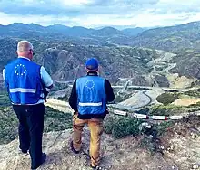 Photographie de deux hommes au bord d'une falaise, observant en contre-bas des camions arrêtés sur une route de montagne.