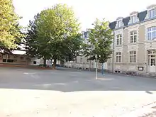 Photographie présentant une cour d'école arborée. Au fond à gauche, un préau. À droite un bâtiment de deux étages (dont un mansardé) sur rez-de-chaussée surélevé, avec un perron de 4 marches devant les portes vitrées.