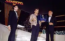 Photographie en couleur de trois personnes sur une scène distribués et équidistants sur une ligne horizontale, la personne au centre tient de sa main gauche un trophée ; en haut à gauche une écriture en lettres majuscules