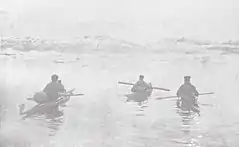 Photographie en noir et blanc de trois hommes chacun embarqué sur un kayak et ramant à proximité d'un littoral.