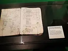 Dans une vitrine, un livret de scénario en mauvais état, ouvert à une page agrémentée de dessins et notes. Des tâches vertes en bas de la page droite.