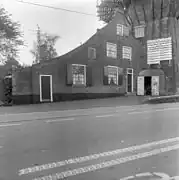 Photographie en noir et blanc d'une maison couronnée d'une galerie et longée par une route.
