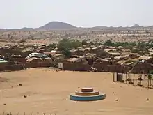 Cliché couleur. Premier plan : étendue de sable. Second plan : derrière une barrière de bambous tentes algnées serrées. Arrière plan : montagnes.