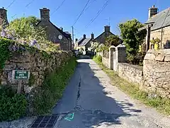Photographie en couleurs prise à l'entrée d'une voie de circulation. Des maisons à gauche et à droite sont séparées de la voie par d'épais murs en pierre.