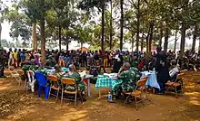 Photographie couleur d'un groupe de militaires en tenue assis derrière des tables, un homme seul également en tenue militaire se trouve face à eux, et derrière se trouve un attroupement de civils.