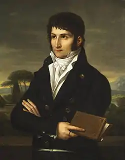 Portrait en buste et les bras croisés de trois quart face à gauche d'un jeune homme d'une trentaine d'années aux cheveux et aux yeux noirs, habillé d'une veste de couleur sombre sur une chemise avec un foulard au cou, tous deux blancs. À l'arrière plan, paysage suggéré de montagne et d'arbres méditerranéens. L'homme tient un livre entrouvert à la main, et regarde hors cadre