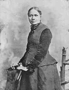Photographie noir et blanc de profil, de trois-quart d'une femme de couleur, cheveux attachés en bandeau, toilette sombre, mains posées sur une chaise