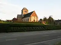 Léger surplomb de l'église.On remarque ses 2 niveaux successifs par rapport à la route. D'abord celui du cimetière, encore entouré de sa haie, auquel on accède par quelques marches.
