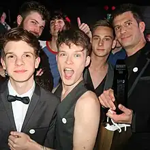 Plusieurs jeunes acteurs sont regroupés et sourient, le contraste est saisissant avec le manager du studio, qui est beaucoup plus âgé et ridé. Le manager tient une récompense dans ses mains.