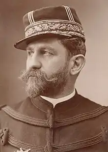 Photo en noir et blanc et de trois-quarts d’un homme en tenue de militaire, cheveux foncés, épaisses moustache et barbe commençant à blanchir, portant une médaille sur le côté droit de la poitrine