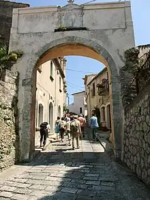 Photographie d'une rue pavée avec au premier-plan une arche voûtée formant une porte d'entrée dans la ville, avec trônant au-dessus une statuette de la Vierge Marie. Un groupe de touristes a passé la porte et monte maintenant dans la rue bordée de maisons anciennes et basses d'un ou deux étages.