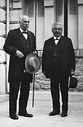 Deux hommes moustachus et vêtus chacun d’un costume foncé sourient sur le perron d'un bâtiment : à gauche, l’homme est chauve, tient dans sa main gauche un chapeau clair et une canne ; à droite, l'individu est plus petit, a les cheveux gris-blancs et tient dans sa main gauche un chapeau de couleur foncée