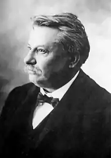 Photographie en noir et blanc ; homme d'une soixantaine d'années légèrement de profil (droit), moustache, costume, nœud papillon