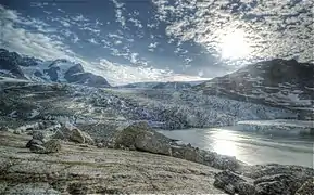 Photographie en couleurs de roches brunes et grises, aux formes arrondies, bordées par les eaux d'un fjord, un glacier visible en arrière-plan.