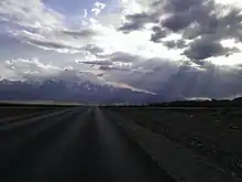 Photographie prise depuis la route, qui n'a pas de marquage. On voit un paysage de la steppe de Kouraï, avec des montagnes aux cimes enneigées au fond. Mais le est ennuagé, les nuages ne laissant que peu passer la lumière, créant une atmosphère sombre.
