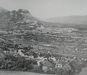 Photographie noir et blanc d'un paysage. Au premier plan se trouve des arbres et quelques bâtiments. Au milieu de l'image un cours d'eau borde une ville. En arrière-plan se trouve un massif montagneux.