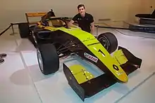 Photographie d'une monoplace de Formule 3 noire et or, vue de trois-quarts dans une exposition, avec un jeune homme à côté.