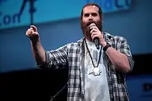 Un homme très barbu, à l'aspect de bûcheron, et portant le logo de l'émission en collier, parle dans un micro face à nous.