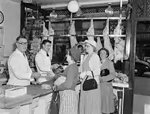 Photo en noir et blanc de l'intérieur d'une boucherie, où on compte deux hommes bouchers et plusieurs femmes clientes.