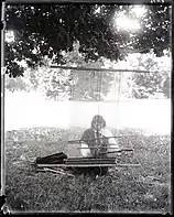 Schwarz-weiß-Foto von We’whas Arbeit an einem Webstuhl. We’wha sitzt dabei auf einer Wiese. We’wha trägt ein bis zu den Füßen reichendes Gewand, dessen Ärmel weiß sind, der Rest ist hingegen schwarz. We’wha trägt eine ähnliche Halskette wie vorher, diesmal stecken in einigen der Steinchen Stacheln. Hinter beziehungsweise über We’wha sind Baumblätter und Bäume zu sehen.