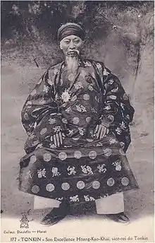 Vieil homme portant un turban, une longue barbe et moustache, une tunique traditionnelle vietnamienne sombre décorée de cercles et de caractères chinois clairs, un pantalon clair et des chaussures sombres. Il est assis.