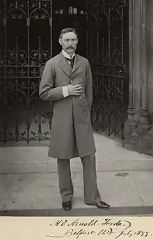 photographie noir et blanc : portrait en pied d'homme moustachu devant une grille de fer forgé