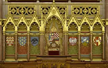 Derrière le pupitre du président de la Chambre des magnats sont représentés les blasons des principales familles ayant régné sur la Hongrie. Encadrant les armes du royaume, de gauche à droite : Arpadiens, Anjou, Hunyadi, Jagellons, Zapolya, Habsbourg.