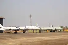 Two four-engine prop-driven aircraft on ramp facing right of screen. The large aircraft in the midground, which is slightly off-centered, has swept-back wings and is painted in a pale grey paints scheme. The other in the background is a straight-wing aircraft painted in pale green