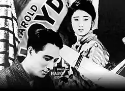 Un homme (Minoru Takada) et une femme (Kinuyo Tanaka) se tiennent devant une affiche où se devine l'inscription en diagonale « Harold Lloyd » (image en noir et blanc).