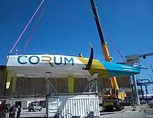 Vue en entier, de profil, en contre-plongée. Suspendu à une grue télescopique, le bateau repose sans mât sur son ber.