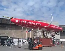 Bateau rouge à foils blancs, sans son mât, posé en hauteur, vu d'en dessous. On voit le haut de la quille, jaune, que des techniciens s'apprêtent à fixer.