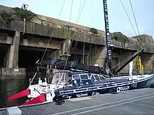 Voilier bleu sombre et blanc, vu de trois quarts arrière, amarré au ponton. En arrière-plan, l'ancienne base de sous-marins allemande de Lorient.
