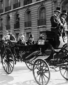 Photo en noir et blanc de deux individus moustachus aux cheveux blancs dans une calèche : l’homme situé à droite porte un haut-de-forme et regarde devant lui ; celui de gauche sourit au photographe ; à l’arrière-plan, deux gardes