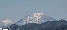 Photo couleur d'un sommet de montagne enneigé sous un ciel bleu avec un massif de collines boisées au premier plan.