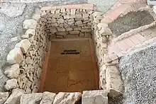 Vue d'une cave aux murs appareillés creusée dans le sol d'une cour antique ; deux canalisations en tuiles aboutissent à la cave.