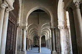 Vue intérieure montrant une double rangée d'arcades en perspective dans le portique précédant la salle de prière. Les arcs sont de type outrepassé (brisé ou en plein cintre).