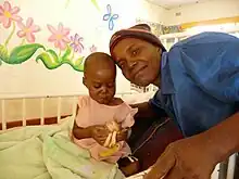 Un nourrisson pose avec un adulte dans une chambre d'hôpital aux murs décorés de peintures florales.