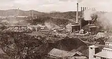 Photographie d'une usine sidérurgique vue de loin. De longues cheminées dépassent d'un corps de bâtiments, et de la vapeur s'échappe de plusieurs endroits.