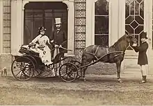 Assis dans une calèche dont le cheval est tenu en mains par un piqueur, Louise et son frère habillés de clair et coiffés d'un chapeau orné posent sous le regard de la future reine Marie-Henriette en tenue luxueuse claire à damiers foncés et du futur roi Léopold&nbsp;II vêtu d'une redingote sombre et d'un pantalon clair et coiffé d'un chapeau haut de forme.