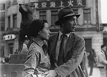 Un homme en costume et chapeau a la main posée sur l'avant bras d'une femme, leurs regards se portent au loin vers la droite. Derrière eux trône la statue d'un chien assi (image en noir et blanc).