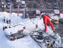 Eva Logar en combinaison rouge avec un dossard, en position de vol au cours d'un saut, vue de trois quarts arrière, paraissant survoler un parking