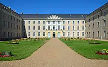 Photographie présentant un grand bâtiment en U à 3 niveaux et toit d'ardoise. Au fond, au bout d'une allée sablée encadrée de pelouses et de massifs de fleurs, l'entrée surmontée d'un fronton sculpté à colonnes.
