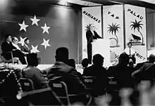 Homme debout parlant derrière un pupitre, devant un drapeau du TTIP occupant la totalité d'un mur